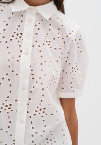 Camicia bianca a maniche corte con motivo a occhio, caratterizzata da ritagli circolari e chiusura con bottoni. Collo fresco e tessuto testurizzato e traspirante.