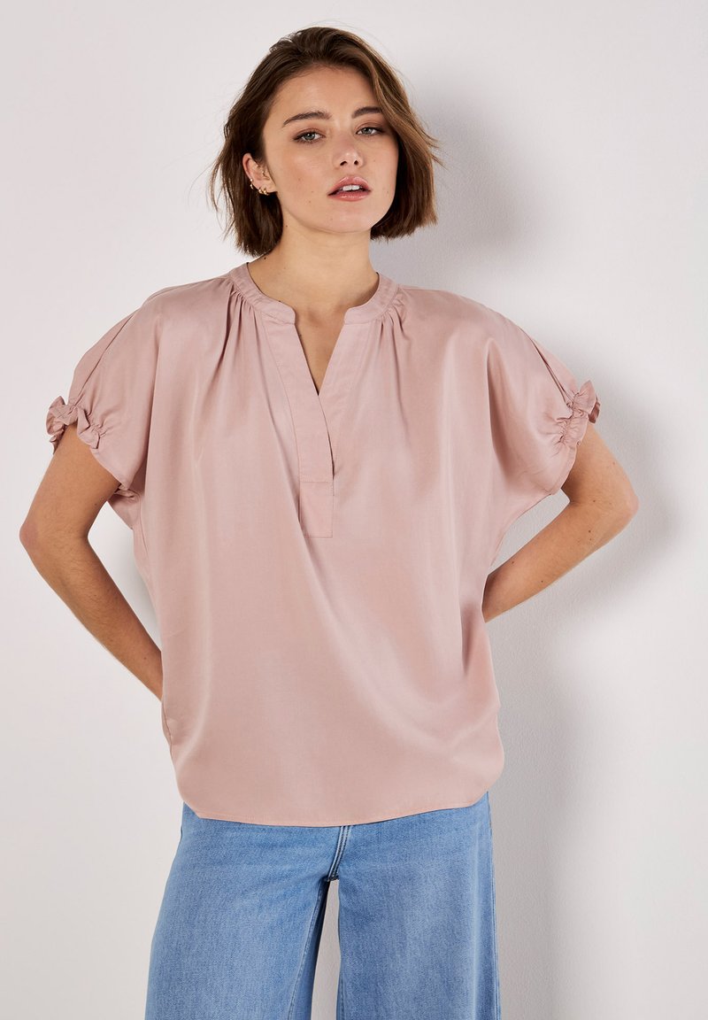 Helle rosa Bluse mit einer lockeren Passform, kurzen Ärmeln mit elastischen Bündchen und einem kleinen V-Ausschnitt. Der Stoff wirkt glatt und leicht.