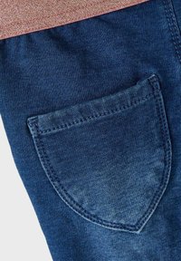 Tummansinisestä denimpuvusta, jossa on läpätaskuyksityiskohta. Saumat ovat kontrastiväriset, ja pinta on sileä, hieman kiiltävä.