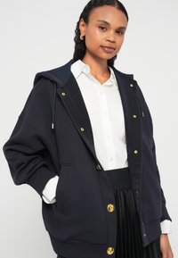 Μπλε σκούρο φούτερ σε μεγάλη διάσταση με χρυσά μεταλλικά στοιχεία, κουκούλα με κορδόνι, τσέπες μπροστά και ριμπ μανσέτες. Συνδυάζεται με λευκό πουκάμισο και μαύρη πλισέ φούστα.