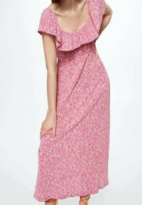 Femme portant une robe midi rose à fleurs avec un col à volants, tenant la jupe d'un côté, debout devant un fond blanc uni.