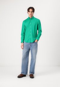 Camicia verde a maniche lunghe con bottoni, abbinata a jeans azzurri chiari e scarpe marroni. La camicia presenta un colletto e un piccolo logo sul petto.