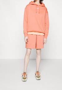 Peach-colored hoodie with drawstrings and Nike logo, paired with matching shorts. Worn with beige and orange sneakers.
