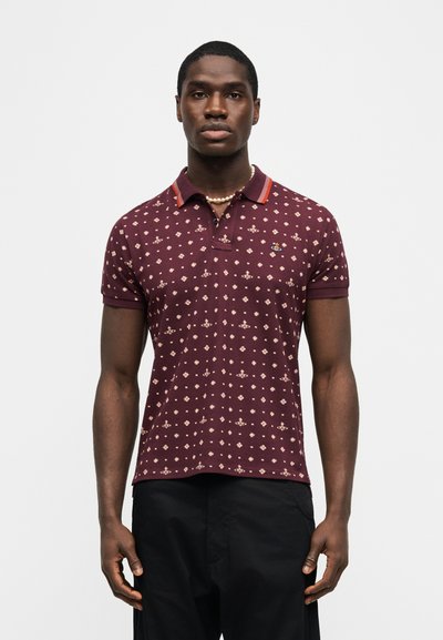 Άνδρας που φοράει μαρουνί πουκάμισο πόλο με κόκκινη ριγωτή γιακά και λευκό κόσμημα με πέρλες, στέκεται μπροστά σε απλό λευκό φόντο.