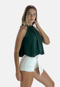 Top verde a collare halter con tessuto drappeggiato, abbinato a pantaloni corti di denim bianchi. Il top presenta una silhouette fluida e una texture morbida.