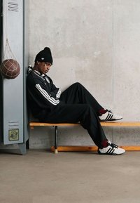 Svart idretts-drakt med hvite striper, båret med en svart lue og hvite joggesko. Trebenk og grå betongvegg i bakgrunnen.