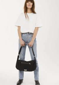 Sac à bandoulière noir en tissu avec une poche avant zippée, montré tenu par une personne portant un t-shirt blanc et un jean bleu clair.