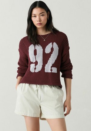 Burgundin villapaita, jossa on suuri valkoinen "92" -grafiikka, ribattu pinta ja alaslasketut olkapäät, yhdistettynä valkoisiin pinstripattuiksi lyhyisiin housuihin.