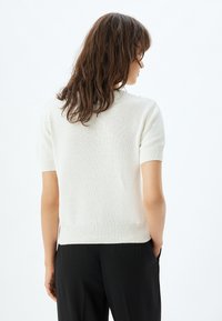 Gebreid T-shirt in lichtcrème, met een geribbelde onderrand en een textuurpatroon, weergegeven vanuit de achterkant tegen een eenvoudige achtergrond.