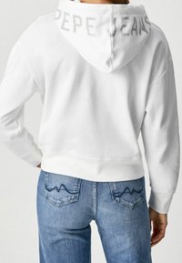 Felpa bianca corta con cappuccio che mostra la scritta "PEPE JEANS" in grigio chiaro. Abbinata a jeans di denim blu con dettaglio di cuciture ondulate sulle tasche.