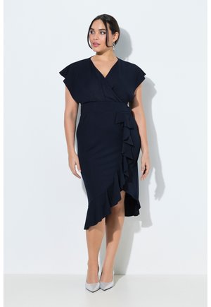 Mujer con un vestido cruzado azul marino con dobladillo asimétrico con volantes y mangas con vuelo, combinado con tacones plateados puntiagudos, sobre fondo blanco.