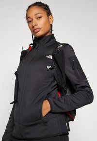 Black fleece jacket with a high collar, featuring zippered pockets and a subtle logo. A black backpack is worn over the jacket.