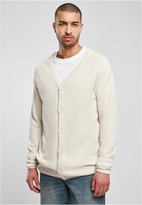 Cremefarbener Cardigan mit V-Ausschnitt, aus strukturiertem Stoff, mit sechs Knopfverschlüssen und gerippten Bündchen, kombiniert mit einem weißen T-Shirt.