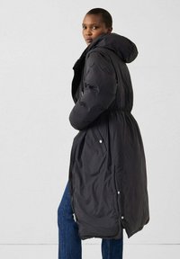 Schwarzer, langer Steppmantel mit Kapuze. Elastisch geraffte Taille, Reißverschluss- und Druckknopfverschluss sowie seitliche Taschen. Glattes Material und isoliertes Design.