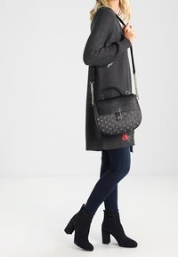 Sac à main noir avec des clous, une poignée supérieure et une bandoulière. Porté avec une robe pull grise, un jean bleu et des bottines noires.