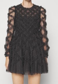 Robe en tulle noir avec des manches longues, présente des volants texturés et un design superposé, des accents transparents avec des détails métalliques subtils.