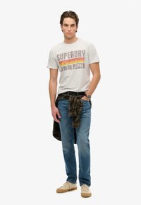 Camiseta gris con estampado "SUPERDRY" en rojo y amarillo, jeans azules y zapatillas beige. Una camisa de cuadros atada a la cintura añade textura.