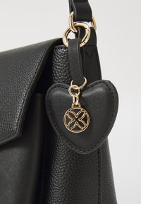Sac en cuir noir avec une finition texturée, présentant un charme en forme de cœur en métal doré et un emblème circulaire décoratif.