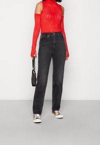 Top largo de manga larga rojo transparente con hombros descubiertos, combinado con jeans negros de talle alto. La modelo sostiene un pequeño bolso negro y lleva zapatillas deportivas coloridas.