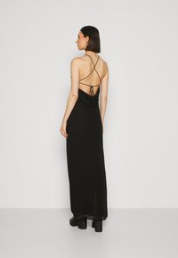 Rochie neagră fără mâneci, cu un design ajustat, bretele crisscross la spate și o textură drapată. Poalele ajung până la glezne, asociată cu tocuri groase.