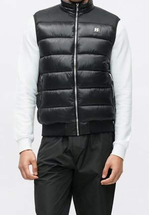 Homme portant une veste sans manches matelassée noire par-dessus une chemise blanche à manches longues et un pantalon noir, debout les bras relâchés le long du corps.