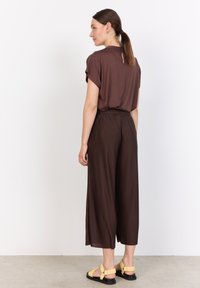 Brown short-sleeve top with back keyhole; matching brown pleated wide-leg pants; black sandals with yellow straps. Minimal design and texture.