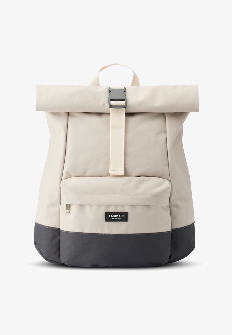 Beige roll-top reppu harmaalla pohjalla, jossa on etutasku, kerma väri olkahihna ja metallinen solki. Brändilabel näkyvillä edessä.