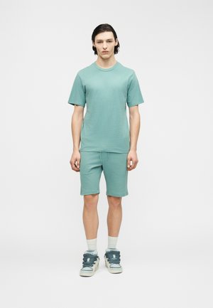 Νεαρός άνδρας στέκεται ανάσκελα φοράει βεραμάν μπλουζάκι με κοντά μανίκια, αντίστοιχα βερμούδα, λευκές κάλτσες και λευκά αθλητικά παπούτσια με βεραμάν κορδόνια.