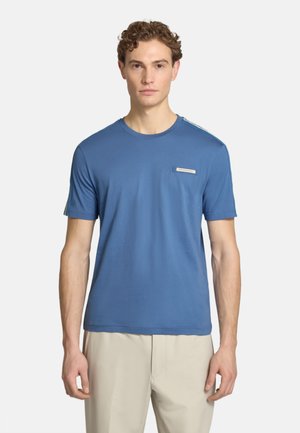 Giovane uomo con capelli corti e ricci che indossa una camicia blu a maniche corte e pantaloni beige chiaro, in piedi davanti a uno sfondo semplice.