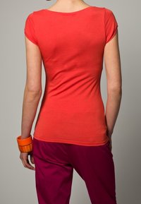 Camiseta ajustada roja de manga corta, con textura suave y escote redondeado, combinada con pantalones rosas y un brazalete naranja en la muñeca.