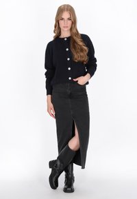 Cardigan blu navy a maglia con cinque bottoni bianchi, abbinato a una gonna di jeans nera con uno spacco frontale. La modella completa il look con stivali neri al ginocchio.