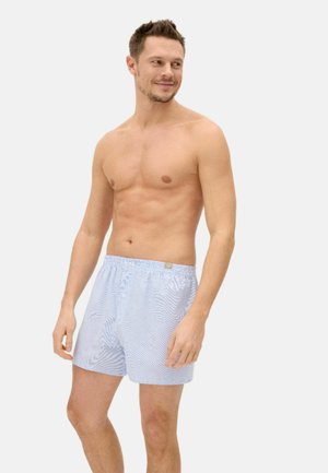 Hombre con bóxers de cuadros azul claro, de pie y sonriendo mientras mira hacia un lado sobre un fondo liso.