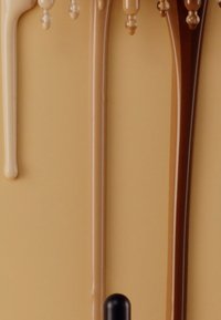 Maquillaje líquido en varias tonalidades gotea de un soporte vertical sobre un fondo beige, exhibiendo texturas suaves y tonos diversos.