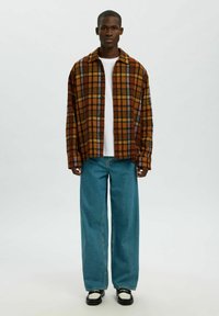 Bruine en oranje geruite overshirt met kraag, witte T-shirt eronder, gecombineerd met losse blauwe jeans en zwarte sneakers.