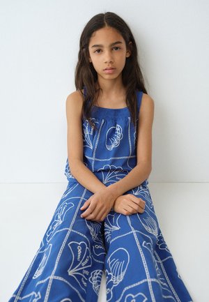 Jeune fille aux longs cheveux foncés assise contre un mur blanc, portant un haut bleu sans manches et un pantalon large assorti avec des motifs de coquillages blancs.