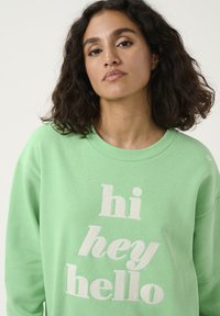 Un sweat-shirt vert clair en tissu doux, orné de texte brodé blanc "hi hey hello". Le col rond et les poignets côtelés sont visibles.