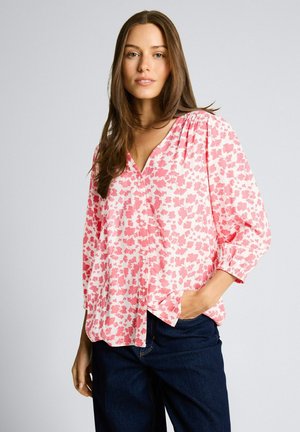 Kobieta o długich brązowych włosach, ubrana w luźną różowo-białą bluzkę w kwiaty oraz ciemnoniebieskie dżinsy, z jedną ręką w kieszeni, na neutralnym tle.