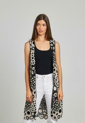 Gilet all'uncinetto bianco e nero con motivi floreali, aperto davanti e senza maniche, indossato sopra una maglietta nera aderente e jeans bianchi.