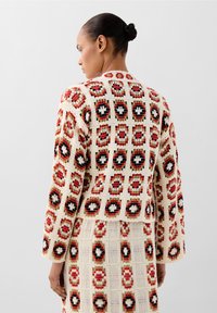 Gehaakte cardigan met een geometrisch patroon van rode, zwarte en crème vierkanten en cirkels. Geweven stof met brede mouwen en een cropped snit.