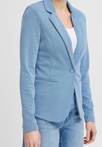 Svetle modrý blejzr z texturované tkaniny, s klopovým límcem, zapínáním na jeden knoflík a dvěma předními kapsami. Vhodný pro smart-casual styl.