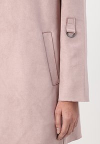 Light pink faux suede coat with a smooth texture, featuring a side pocket and a strap detail with a metal ring on the sleeve.