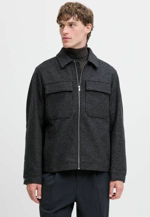 Young man wearing a dark gray zip-up jacket with two chest pockets over a black turtleneck, looking to the side.