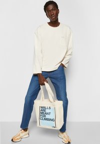 Beige langærmet skjorte, blå jeans og en cremefarvet tote taske med teksten "VÆGGE ER TIL AT KLATRE" i sort og blå. Sneakers med orange detaljer.