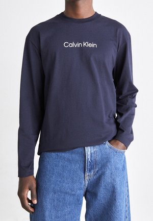 Persona che indossa una maglia a maniche lunghe Calvin Klein blu navy infilata in un paio di jeans blu a vita alta, con una mano in tasca.
