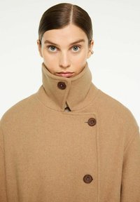 Beige uldfrakke med høj krave, fastgjort med to brune knapper. Stoffets tekstur er blød, og designet er oversized.