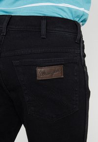 Close-up of black denim jeans back pocket with brown leather Wrangler brand label, worn with a teal and white striped shirt.