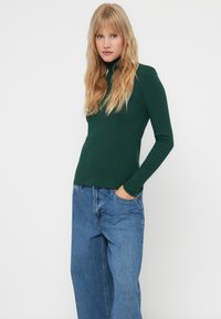 Zeleni dugi rukav turtleneck top, rebrasta tekstura, prilagođeni dizajn. Uparen s lagano širokim svijetloplavim trapericama.