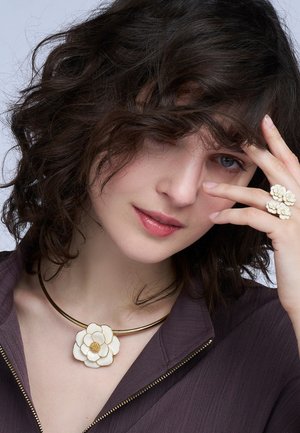 Collier ras du cou doré avec un pendentif en forme de fleur blanche, associé à des bagues florales assorties. Des cheveux bouclés et un haut violet profond sont visibles.