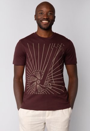 Bordeaux kleurig T-shirt met korte mouwen met een beige grafiek van een wiel en lijnen die naar buiten stralen. Zacht katoenen materiaal met een ronde hals.