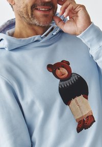 Sudadera azul claro con un gráfico de un oso que lleva un suéter negro y pantalones beige, con detalles en calzado naranja y negro.